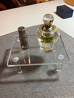 Two perfume bottles displayed on a clear acrylic stand: a small sterling silver bottle with screw-on lid and a larger cut crystal bottle with stopper, placed on a textured surface.