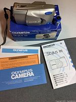 Silver Olympus Stylus 105 camera on original blue box with instruction manuals and Energizer battery placed in front on grey surface.
