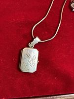 Rectangular vintage sterling silver locket with floral engraving and attached chain on red fabric background, showing front and clasp side