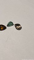 Three pear-cut natural rainbow tourmaline gemstones in orange, teal, and light purple champagne colors placed on a white surface.