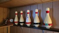 Five vintage bowling pins with red and black detailing, displayed on a wooden shelf against wood paneling.