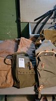 Wide shot showing multiple canvas bags in tan, olive, and beige colors arranged on a table with visible wear and hardware