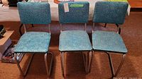 Three blue vinyl retro chairs with chrome metal tubular legs and sled bases, legs with rust marks, arranged in a basement room with brown carpet and wood-paneled walls.