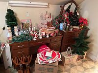 Wide view of Christmas decorations arranged on two wooden dressers showing trees, figurines, electric candles, and boxed items including a 2.5-foot artificial tree.