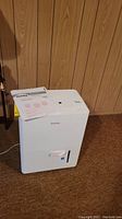 White Danby dehumidifier with user manual placed on top, showing front view in basement setting.