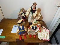 Four vintage collectible dolls arranged on a wooden surface, two on display stands, one with folk-style clothing including apron and headscarf.