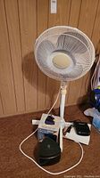 White standing floor fan, black triangular portable heater, and black car heater placed on carpeted floor against wood-paneled wall.