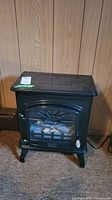Front view of small black electric faux fireplace heater with glass panel and faux logs inside, sitting on carpet with wood-paneled wall behind.