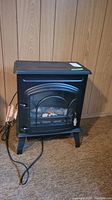 Front view of compact black electric faux fireplace heater standing on carpet against wood panel wall, showing power cord and front glass door with faux logs inside.