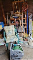 Photo of garden chair, hose reel, and a variety of wooden-handled garden tools leaning against a wooden wall.