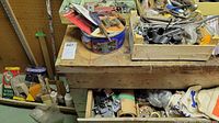 Photo showing a wooden workbench with an open drawer containing assorted hardware pieces, tins, and tools, along with canvas aprons and metal oil cans placed nearby.