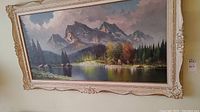 View of entire framed landscape painting hanging on wall showing mountain, lake and forest scenery.