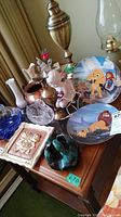 Full view of table displaying all items in lot with oil lamp, Disney plates, swan figurines, pottery dish, and vases.