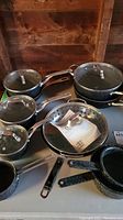 Photo showing a collection of black speckled cookware including three saucepans with lids, a frying pan, a large pot with lid, and metal strainer insert on a table.