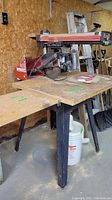 Side-angle photo of Craftsman radial arm saw mounted on a wooden work table with dust collection bucket underneath, showing saw blade, handle, and table surface.