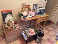 Wide view showing all sewing items on and around a wooden table and small stand, including threads, scissors, patterns, trash cans, and a small wooden cabinet.