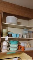 Three shelves of assorted Tupperware and food storage containers, including large white container, turquoise bowls, beige bowls, orange cups, citrus reamers, and measuring devices.