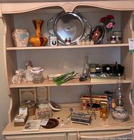 Three shelves containing glass and ceramic vases, metal tray, watches, oil lamp, trinket boxes, candles, rosary and other decorative items.