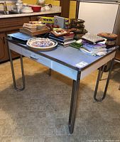 Photo of vintage laminate top table with metal frame, wood drawer and hairpin legs, loaded with various household items.
