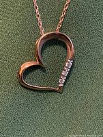 Front view of rose gold heart pendant with five small diamonds on one edge, on a rose gold chain, set against green fabric.