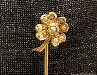 Close-up of 14k gold floral stick pin showing floral design with pearls on each petal and the stem, placed on black textured background.
