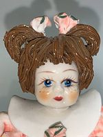 Close-up of one porcelain figurine girl's face showing brown hair styled in pigtails with white and pink flower decorations, blue eyes, and rosy cheeks.