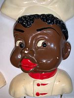 Close-up of one chalkware chef figure showing dark brown skin, black hair, red lips, and white chef uniform with red details.