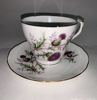 Front view of the bone china teacup sitting on matching saucer showing purple thistle decoration and gold trim