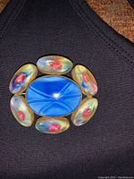 Front view on black fabric showing blue central glass stone surrounded by six multicolored oval glass stones in brass.