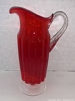 Front and side view of large red glass pitcher with clear applied handle showing ribbed vertical design and glossy finish
