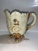 Front and side view showing scalloped rim, embossed floral and scroll design in milk glass with gold paint, resting on curled gold feet.