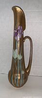 Front view of tall porcelain ewer showing hand-painted purple and white iris flowers on a gold-tone background with elegant curved spout and handle.