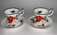 Two Royal Albert Yuletide pattern teacups and matching saucers side by side, showing detailed festive red poinsettia and holly berry decoration with gold trim.