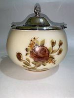 Front view of biscuit jar showing floral design in brown and rose colors on cream milk glass, with silver lid topped by finial.