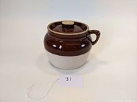 Front view of vintage bean pot showing rounded brown glazed upper part, beige bottom, lid with knob, and handle.