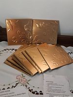 Gold coloured aluminum decorative tiles with embossed floral pattern, arranged stacked with two tiles standing upright behind them.