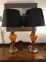 Pair of amber glass and brass lamps with black shades placed on wooden surface in front of a decorative wall mirror.