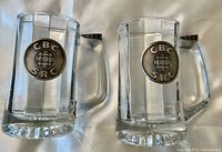 Front view of both glass tankards with CBC SRC metal circular logos and metal thumb handles.