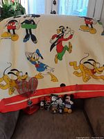 The Disney collectible lot arranged on a sofa including a cream and red colored blanket featuring Mickey Mouse, Goofy, Donald Duck, Pluto, along with Mickey themed shampoo, hand soap, salt and pepper shakers, a small Mickey Mouse plush, and a red metal heart-shaped LOVE trinket dish.