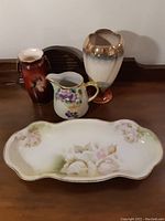 Lot overview with all four pieces on wooden surface: portrait vase, floral pitcher, gold-accented tall vase, and floral tray
