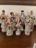 Front view of seven porcelain figurines on a wooden surface beneath a framed picture, showing their detailed historical costumes and poses.