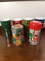 Photo showing six M&M plastic cups, a holiday-themed tin with colorful M&M characters, and a red and white M&M thermos with two characters on the side.