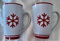 Photo showing two tall ceramic mugs with red snowflake design and red rim stripes, side view.