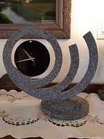 Full view of the modern spiral shaped clock on a table with intricate granite textured frame and black circular dial.