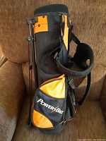 Full side view of the Power Bilt child golf bag showing the orange and black color scheme, shoulder harness, and zippered pocket with logo.