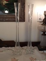 Full view of the pair of tall clear glass vases, showing slender neck and wide 4-legged base.