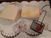 Four items arranged on a white cloth with floral embroidery: ceramic paper bag-shaped dish, small hand-painted ceramic dish, wire basket with fruit decoration, white marble lidded box.