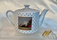 Front side view of white ceramic teapot decorated with brown paw prints and a framed cat painting on the body, with rubber pouring spout placed beside it.