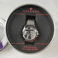 Close-up image of the Oscardo Inukshuk watch in its round metal case, showing the silver-tone watch with mesh metal band and Inukshuk design on dial.