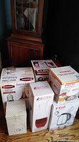 Photo shows multiple boxed and unpacked appliances including TFal kettle, Starfrit food processor, Bravetti wine chiller, and Cuisinart frozen yogurt maker, along with glass carafe and pitcher on wooden floor.